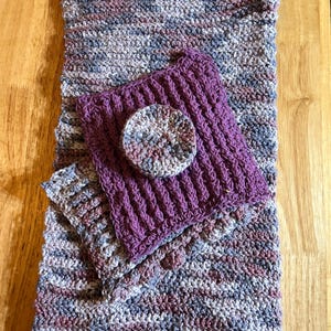 May include: Three crocheted squares in shades of purple, gray, and pink. The largest square is a striped pattern, the medium square is a solid purple, and the smallest square is a striped pattern.