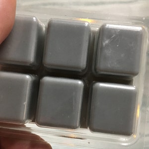 Puede incluir: Primer plano de un recipiente de pl&aacute;stico transparente que contiene seis cubos de cera gris. Los cubos son cuadrados con bordes redondeados y una superficie lisa. El recipiente est&aacute; sujeto por los dedos de una persona.