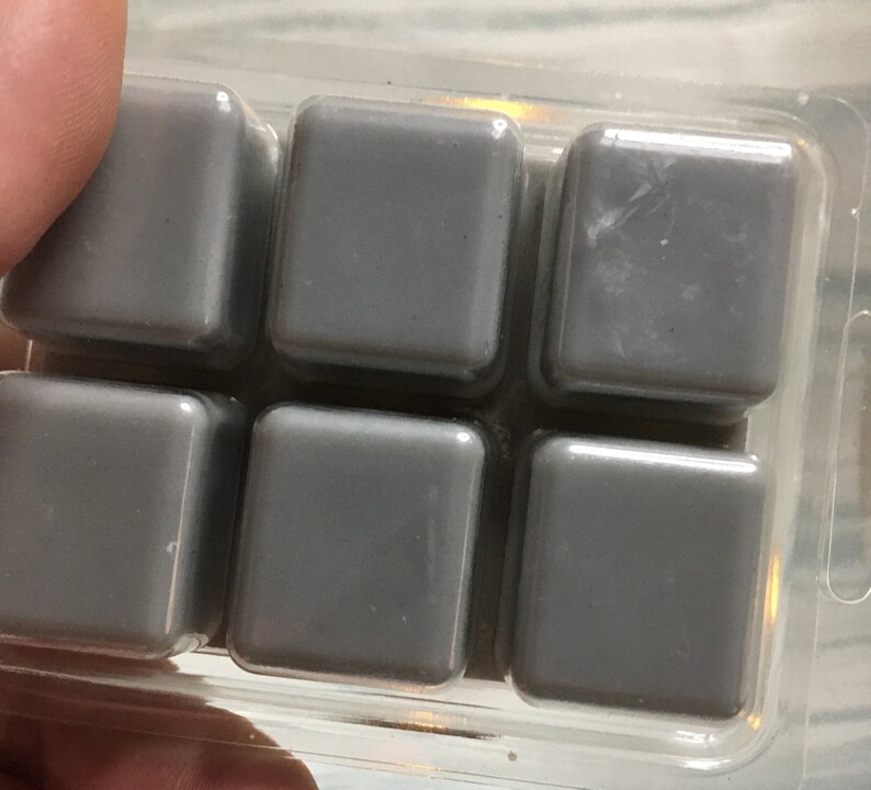 Puede incluir: Primer plano de un recipiente de pl&aacute;stico transparente que contiene seis cubos de cera gris. Los cubos son cuadrados con bordes redondeados y una superficie lisa. El recipiente est&aacute; sujeto por los dedos de una persona.