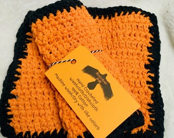May include: Two orange crocheted dishcloths with black borders. The dishcloths are stacked on top of each other. A yellow tag with black text reads "Hand Crocheted Washcloth/Dishcloth 100% Cotton Machine Wash/Dry with like colors".