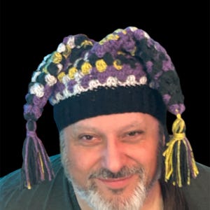 May include: A handmade crochet hat with a black brim and a colorful top. The top features a pattern of purple, yellow, black, and white squares. The hat has two long, dangling tassels with purple and yellow strands.
