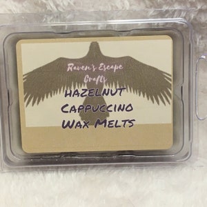 Op de afbeelding: Een doorzichtige plastic container met een label waarop staat "Raven's Escape Crafts Hazelnut Cappuccino Wax Melts". Het label toont een bruin silhouet van een vogel met uitgespreide vleugels.