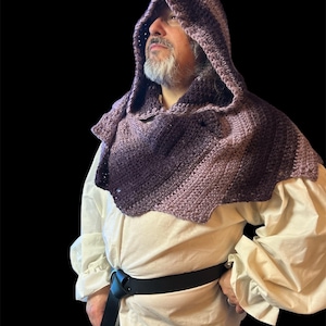May include: A crocheted hooded cowl in shades of purple and lavender. The cowl is worn over a white shirt with puffy sleeves and a black belt. The background is black.