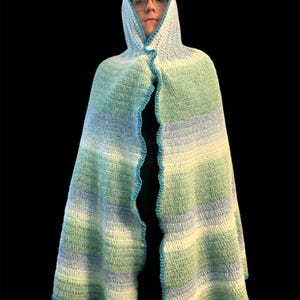 May include: A hooded cape in shades of green, blue, and white, with a scalloped turquoise trim. The cape is crocheted and has a button closure at the neck. The garment is designed to be worn.