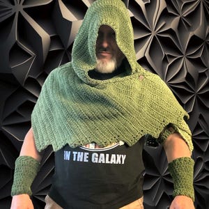 May include: A green hooded cloak, matching wrist cuffs, and a black t-shirt with the text "IN THE GALAXY". The cloak and cuffs are made of a textured, knitted material. The background is a dark, geometric pattern.