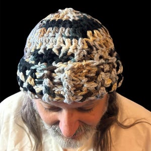 Könnte beinhalten: Ein handgehäkelter Hut in einer Mischung aus Farben, darunter Schwarz, Grau, Beige und Türkis. Der Hut ist mit einem strukturierten Stich gefertigt und hat eine abgerundete Krone. Der Hut ist ein Winteraccessoire.