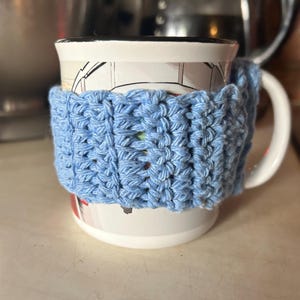 Può includere: Una tazza in ceramica bianca con un rivestimento in crochet blu. La tazza ha un disegno di un autobus a due piani rosso.