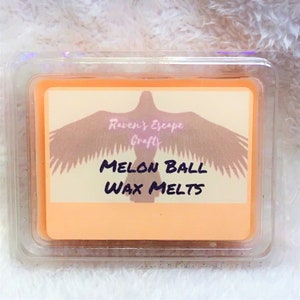 Könnte beinhalten: Ein quadratischer, transparenter Plastikbehälter mit einem pfirsichfarbenen Wachs-Schmelzstück darin. Das Etikett auf dem Wachs-Schmelzstück lautet "Raven's Escape Crafts" und "MELON BALL WAX MELTS".