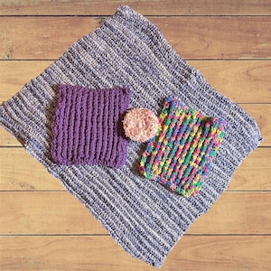 Pode incluir: Três panos de cozinha de crochê, um roxo, um multicolorido e um rosa, sobre uma superfície de madeira.