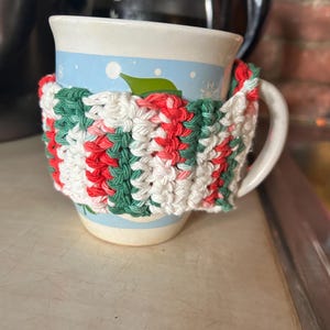 Op de afbeelding: Een witte keramische mok met een blauw en groen wintertafereel. De mok is gewikkeld in een gehaakte cozy met rode, witte en groene strepen.