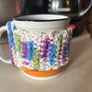 Op de afbeelding: Een witte keramische mok met een zwarte rand en een gehaakte cozy in tinten paars, groen, blauw en wit. De cozy is gemaakt met een granny square patroon.