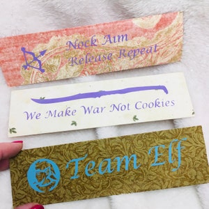 May include: Three rectangular decorative signs with text and graphics. The top sign is pink with gold accents and the text "Nock Aim Release Repeat". The middle sign is white with a purple sword graphic and the text "We Make War Not Cookies". The bottom sign is green with the text "Team Elf".