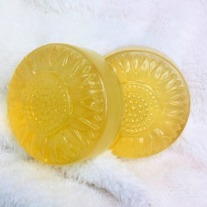 May include: Two round yellow soap bars with a sunflower design. The soap bars are on a white fluffy surface.