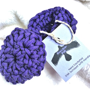 Puede incluir: Dos estropajos redondos, tejidos a mano, en un vibrante púrpura con brillo metálico. Una etiqueta con un gráfico de cuervo y el texto "Hand Crocheted Dish Scrubber With Handle" está adjunta. Los estropajos están sobre una superficie blanca y esponjosa.