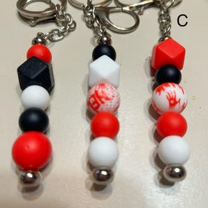 May include: Three keychains with beaded accents in red, black, and white. Each keychain features a different geometric bead, including a cube, a die, and a round bead with a red and white pattern. The keychains have silver-colored hardware.