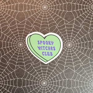 Op de afbeelding: Een groene sticker in de vorm van een hart met een witte rand. De sticker heeft de tekst "SPOOKY WITCHES CLUB" in paarse letters.
