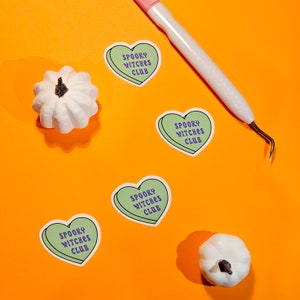 Op de afbeelding: Vier groene hartvormige stickers met de tekst "Spooky Witches Club" in paarse letters op een feloranje achtergrond. Twee witte decoratieve pompoenen bevinden zich ook op de achtergrond.