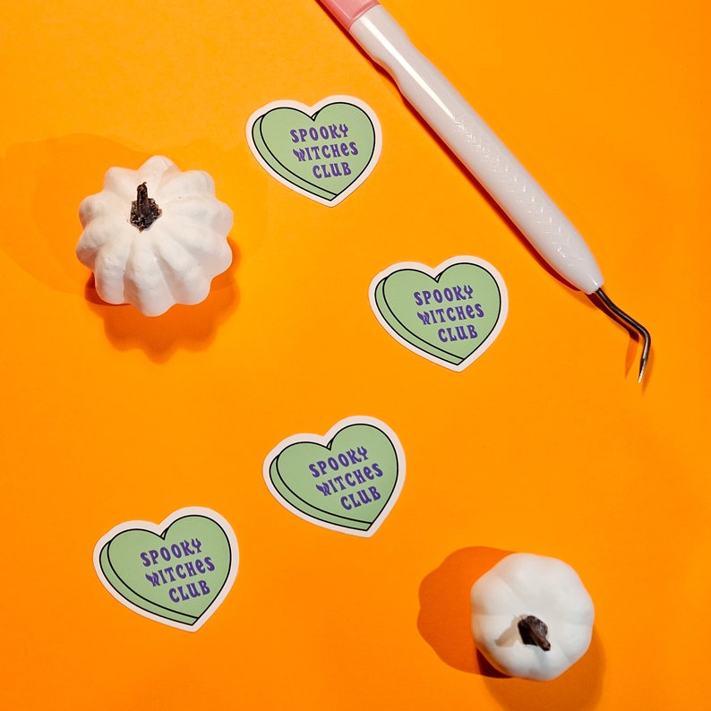 Op de afbeelding: Vier groene hartvormige stickers met de tekst "Spooky Witches Club" in paarse letters op een feloranje achtergrond. Twee witte decoratieve pompoenen bevinden zich ook op de achtergrond.