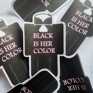 May include: Black and white sticker with the text "Black is her color" in pink letters. The sticker is shaped like a person wearing a dress.