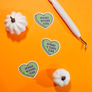 Op de afbeelding: Drie groene hartvormige stickers met de tekst "Spooky Witches Club" in paarse en oranje letters op een oranje achtergrond. Er zijn twee witte pompoenen op de achtergrond.