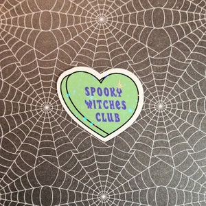 Op de afbeelding: Een groene sticker in de vorm van een hart met glitteraccenten. De sticker zegt "Spooky Witches Club" in paarse letters.