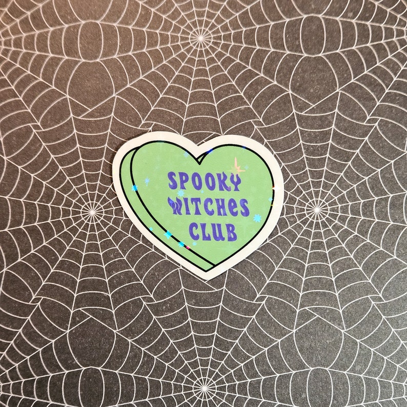 Op de afbeelding: Een groene sticker in de vorm van een hart met glitteraccenten. De sticker zegt "Spooky Witches Club" in paarse letters.