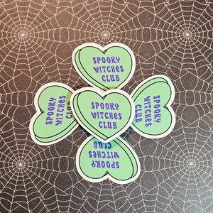 Op de afbeelding: Vier groene hartvormige stickers met de tekst "Spooky Witches Club" in witte en paarse letters.