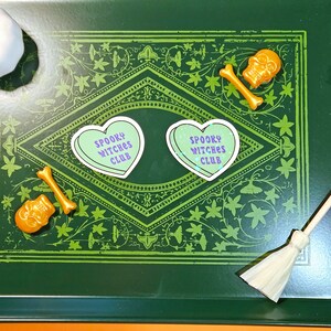 Op de afbeelding: Twee groene hartvormige stickers met de tekst "Spooky Witches Club" in paarse letters op een groene achtergrond met een decoratieve rand. Er zijn twee gouden schedel- en beenderdecoraties en een kleine bezem.