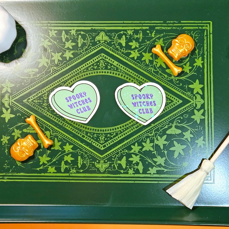 Op de afbeelding: Twee groene hartvormige stickers met de tekst "Spooky Witches Club" in paarse letters op een groene achtergrond met een decoratieve rand. Er zijn twee gouden schedel- en beenderdecoraties en een kleine bezem.