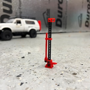 Könnte beinhalten: Ein roter und schwarzer Miniatur-High-Lift-Wagenheber, ein Werkzeug zum Anheben von Fahrzeugen. Der Wagenheber befindet sich auf einer Betonoberfläche, mit einem weißen SUV im Hintergrund. Der Wagenheber ist etwa 10 cm hoch.