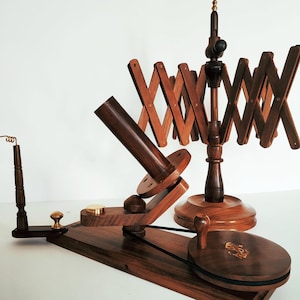 May include: A wooden yarn swift with a folding design and a spindle for winding yarn. The swift has a dark brown finish and a brass knob. The spindle is attached to a wooden base with a black rubber band.