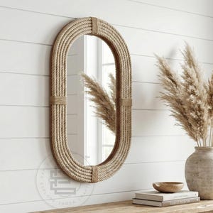 May include: Oval-shaped mirror with a rope-wrapped frame. The mirror is mounted on a white wall with horizontal paneling. The frame is made of natural-colored rope, and the mirror reflects the room. A vase with dried plants sits on a wooden surface.
