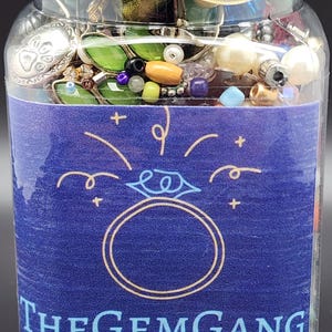May include: A clear jar filled with various beads and jewelry components. The jar has a blue lid tied with a yellow ribbon. A blue label on the jar reads "THEGEMGANG" with a ring graphic.