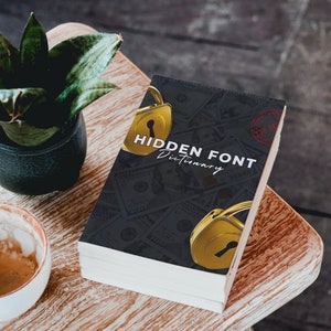 May include: A black and white book with the title "HIDDEN FONT Dictionary" on the cover. The cover features a gold padlock with a keyhole and a red stamp that says "TOP SECRET". The book is sitting on a wooden surface.