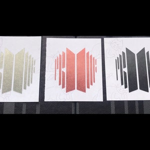 May include: Three versions of the same logo, one in silver, one in rose gold, and one in black. The logo is a stylized representation of the word "BTS" in a geometric font.