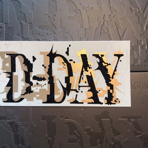 May include: A white sticker with black and gold lettering that reads "D-DAY" in a stylized font. The sticker is on a gray background with the text "August D" in the bottom right corner.