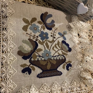 Op de afbeelding: Een beige linnen kussen met een geborduurd bloemenmotief met een vaas met blauwe en bruine bloemen. Het kussen is afgewerkt met witte kant.
