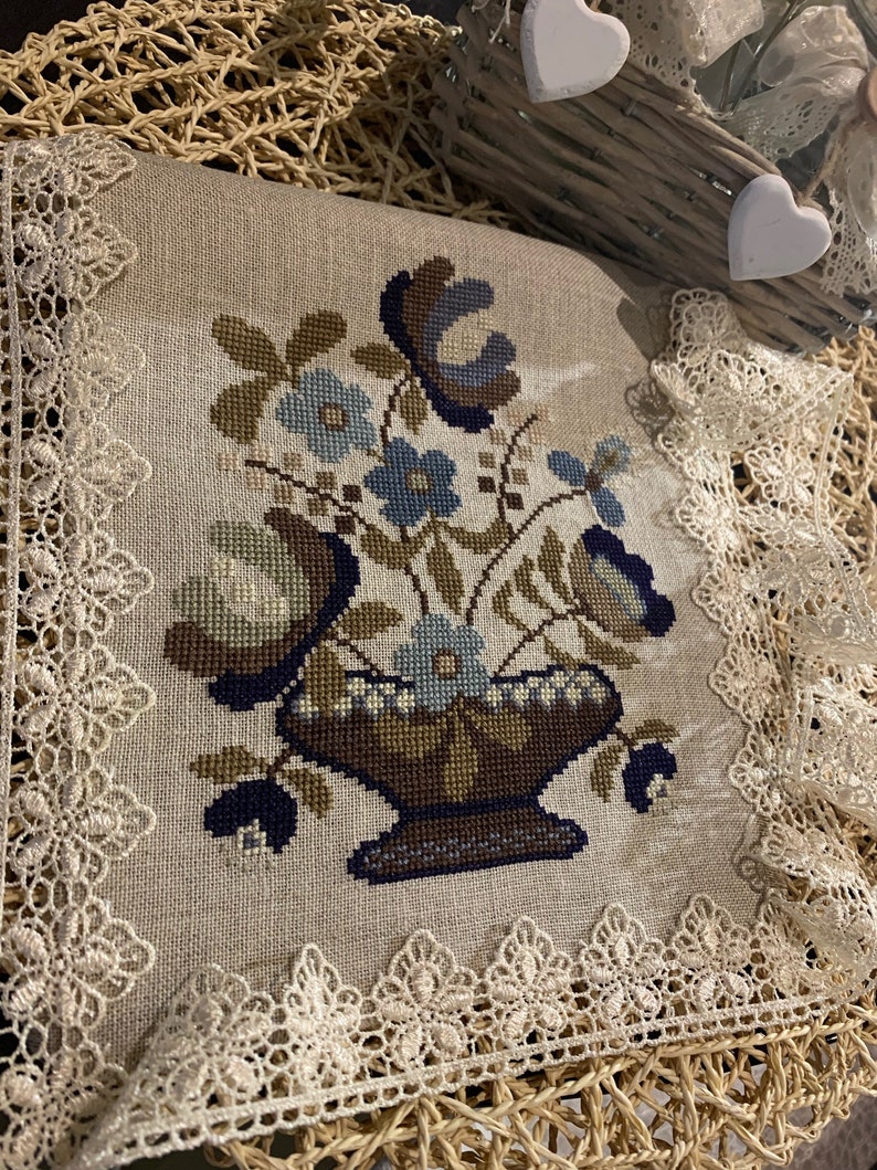 Op de afbeelding: Een beige linnen kussen met een geborduurd bloemenmotief met een vaas met blauwe en bruine bloemen. Het kussen is afgewerkt met witte kant.