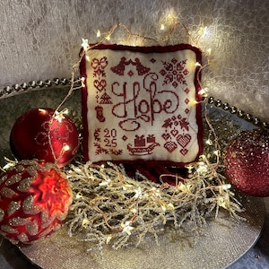 Könnte beinhalten: Ein weißes und rotes Kreuzstich-Kissen mit dem Wort "Hope" in rot gestickt. Das Kissen ist von einer Lichterkette und roten Weihnachtskugeln umgeben.
