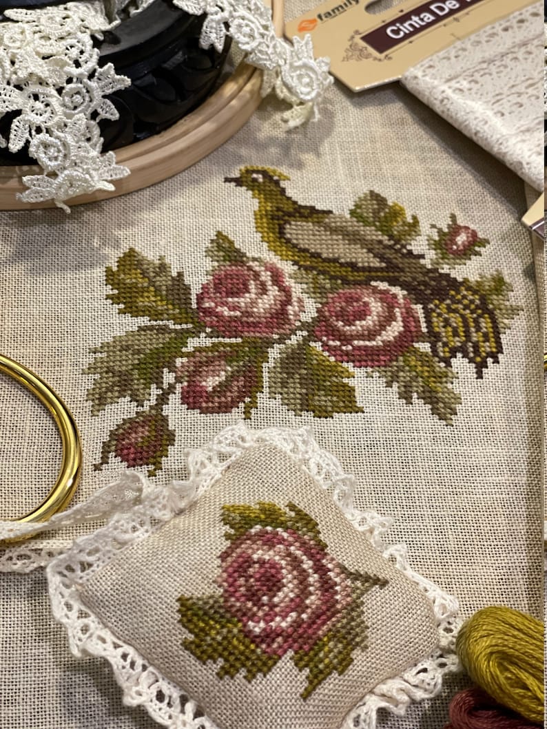 K&ouml;nnte beinhalten: Ein Kreuzstich-Stickprojekt mit einem Vogel, der auf einem Zweig mit rosa und wei&szlig;en Rosen sitzt. Das Projekt ist gerahmt und auf einem beigefarbenen Leinenstoff ausgestellt. Ein kleines Kissen mit einem &auml;hnlichen Rosendesign ist ebenfalls abgebildet.