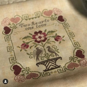 Könnte beinhalten: Ein Kreuzstich-Stickmuster mit zwei Vögeln in einer Vase mit Blumen und dem Text "Two hearts one love". Das Muster ist von einem Rand mit Herzen und einem geometrischen Muster umgeben.