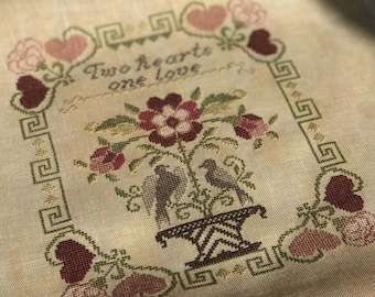 May include: A cross-stitch embroidery design featuring two birds in a vase with flowers and the text "Two hearts one love". The design is surrounded by a border of hearts and a geometric pattern.