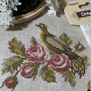 K&ouml;nnte beinhalten: Kreuzstichmuster eines Vogels, der auf einem Zweig mit rosa Rosen sitzt. Das Muster ist auf einem beigefarbenen Aida-Stoff und befindet sich in einem Holzrahmen.