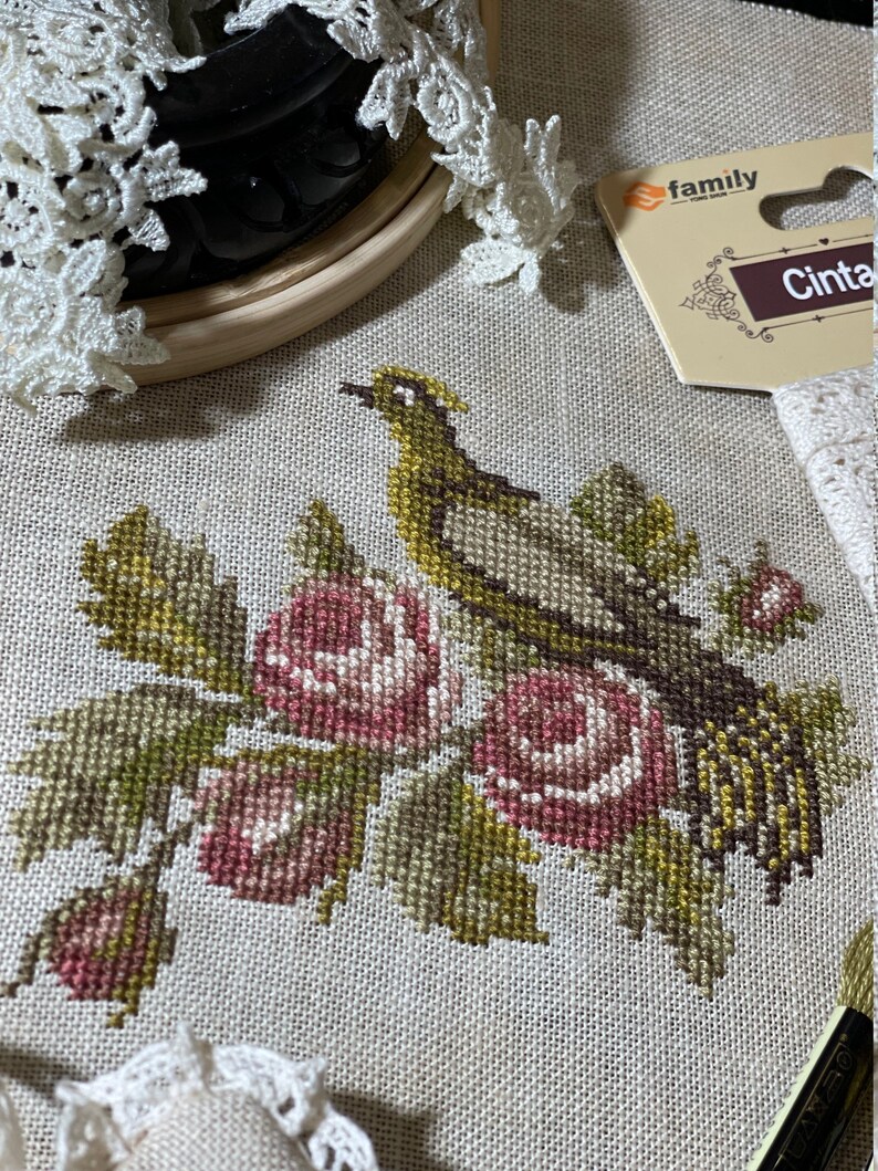 K&ouml;nnte beinhalten: Kreuzstichmuster eines Vogels, der auf einem Zweig mit rosa Rosen sitzt. Das Muster ist auf einem beigefarbenen Aida-Stoff und befindet sich in einem Holzrahmen.