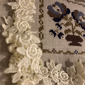 K&ouml;nnte beinhalten: Ein wei&szlig;er Spitzenbesatz mit floralem Muster, der eine Kreuzstich-Stickerei eines Blumenstrau&szlig;es in Blaut&ouml;nen, Braun und Gr&uuml;n einrahmt.