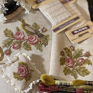 K&ouml;nnte beinhalten: Ein Kreuzstichprojekt mit einem Vogel, der auf einem Rosenbusch sitzt. Das Projekt ist teilweise fertiggestellt und umfasst Stickgarn in den Farben Gr&uuml;n, Rosa und Braun. Das Projekt wird auf einem beigefarbenen Leinenstoff mit einer Spitzenborte pr&auml;sentiert.