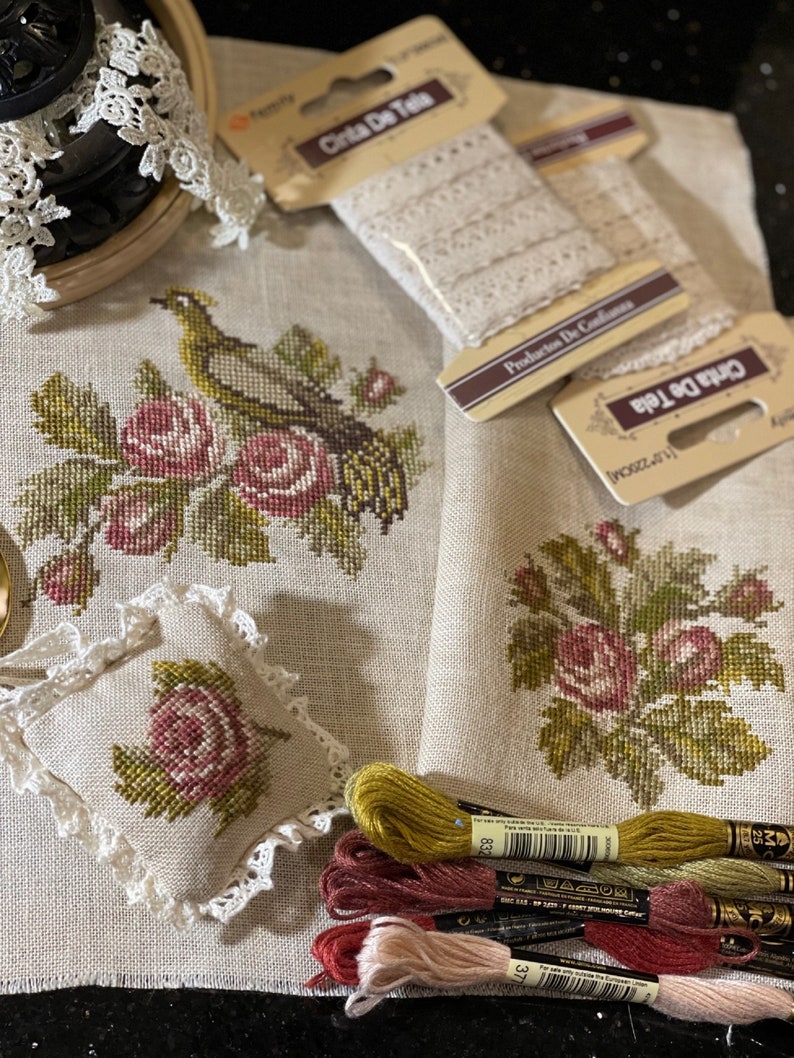 K&ouml;nnte beinhalten: Ein Kreuzstichprojekt mit einem Vogel, der auf einem Rosenbusch sitzt. Das Projekt ist teilweise fertiggestellt und umfasst Stickgarn in den Farben Gr&uuml;n, Rosa und Braun. Das Projekt wird auf einem beigefarbenen Leinenstoff mit einer Spitzenborte pr&auml;sentiert.