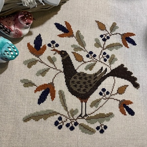 May include: A cross-stitch embroidery of a brown bird perched on a branch with green leaves and orange and blue flowers. The bird is surrounded by small blue berries.