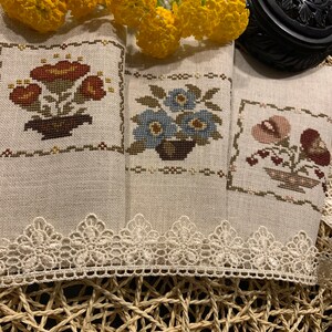 K&ouml;nnte beinhalten: Drei bestickte Geschirrt&uuml;cher mit Blumen- und Pilzmotiven. Die Handt&uuml;cher sind beige mit einer wei&szlig;en Spitzenborte und liegen auf einer geflochtenen Unterlage.