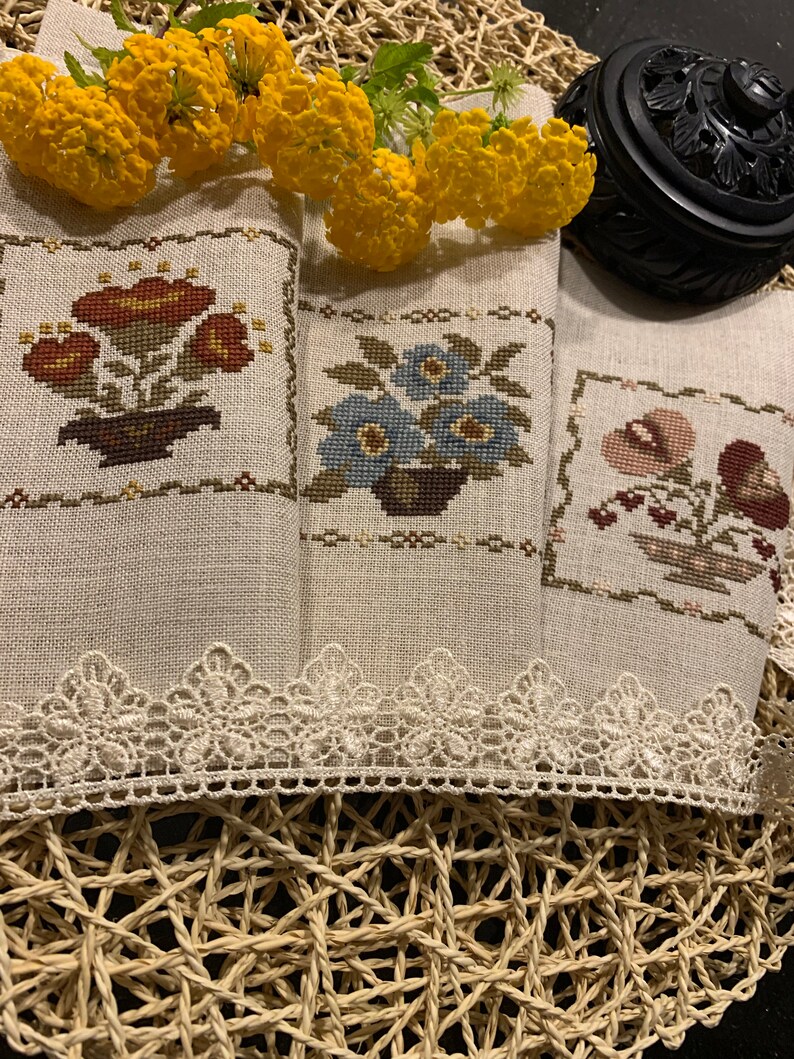 K&ouml;nnte beinhalten: Drei bestickte Geschirrt&uuml;cher mit Blumen- und Pilzmotiven. Die Handt&uuml;cher sind beige mit einer wei&szlig;en Spitzenborte und liegen auf einer geflochtenen Unterlage.
